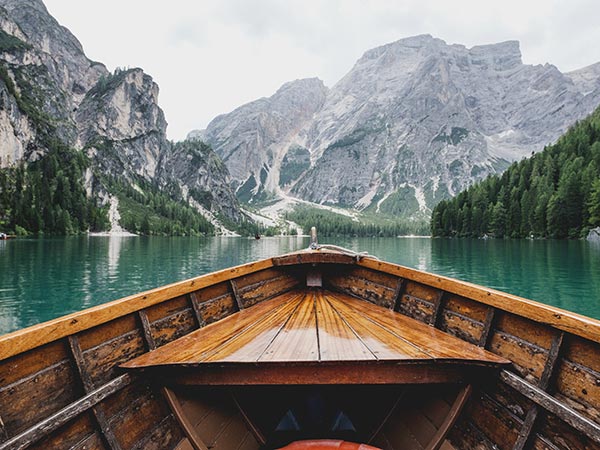 A wooden boat's bow overlooks a tranquil mountain lake, surrounded by towering cliffs and lush forest, under a cloudy sky.