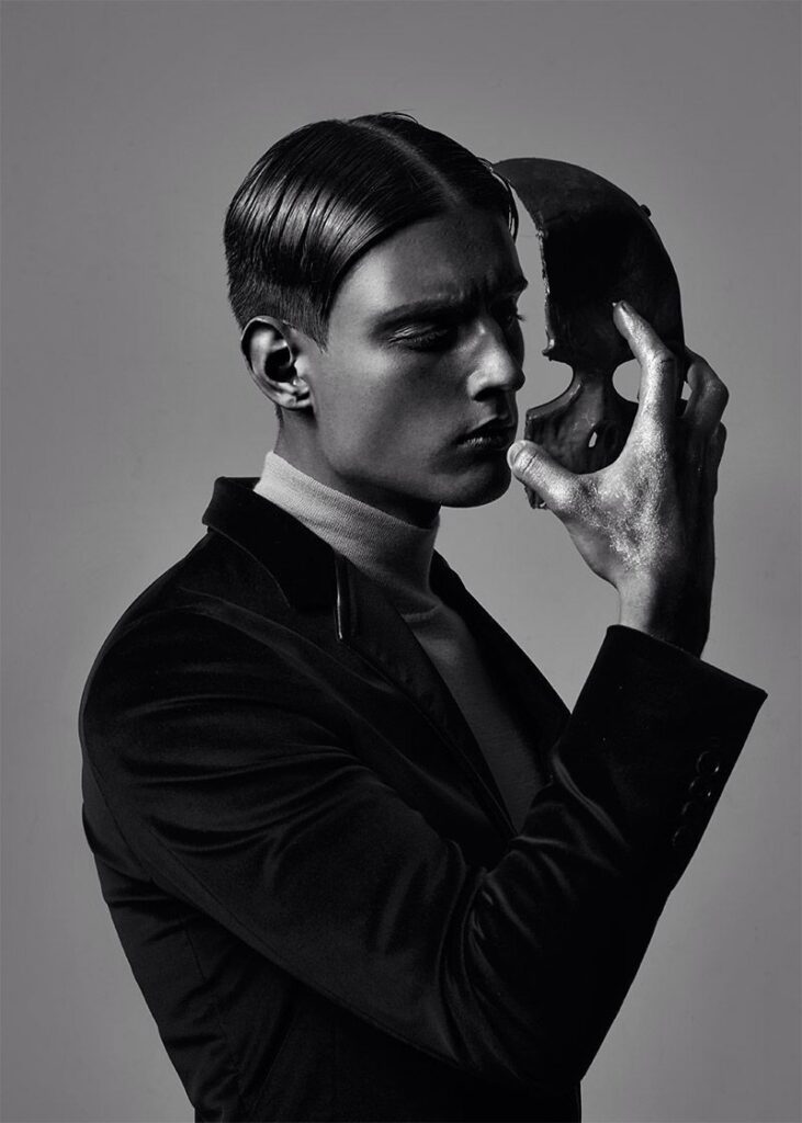 A man in a black suit and turtleneck examines a dark mask, reflecting a mood of introspection and mystery.