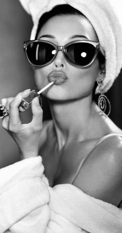 A woman with oversized sunglasses and a towel on her head applying lip gloss while wearing a plush robe.
